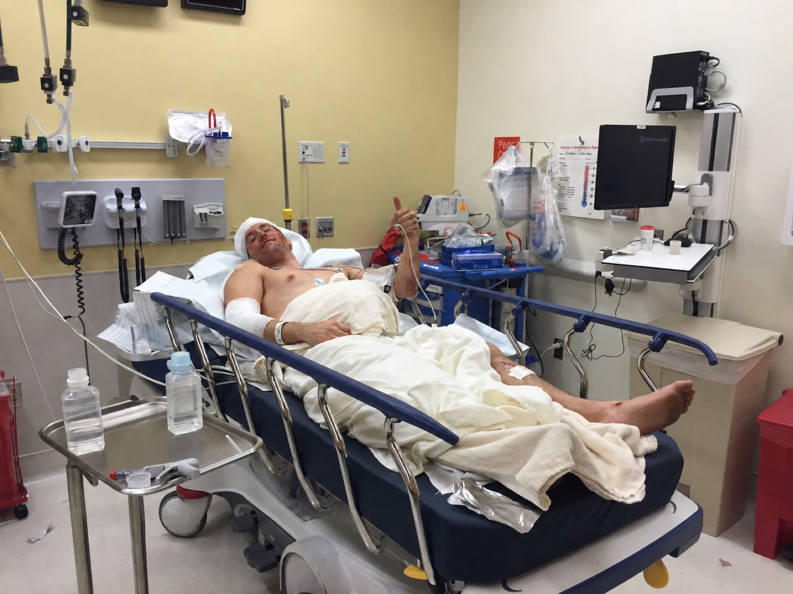 Boyd Johnson on a gurney in the emergency department, head and arm bandaged, giving a thumbs up